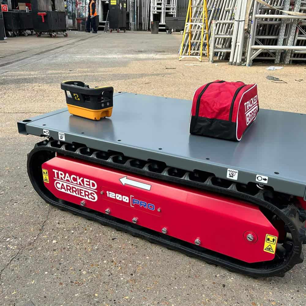 A red and gray Tracked Carriers 1200 Pro 4 crawler platform sits on pavement, with a remote control and a red bag placed on top.